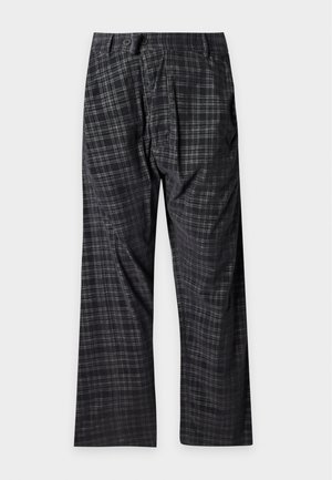 Schwarzer männlicher Model trägt ein transparentes schwarzes Langarmhemd, schwarze Karohose mit weitem Bein und dunkelbraune und beige Loafers, steht vor weißem Hintergrund.