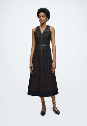 Femme aux cheveux courts et bouclés porte une robe midi noire sans manches avec poches avant et chaussures plates noires à lacets sur fond uni.