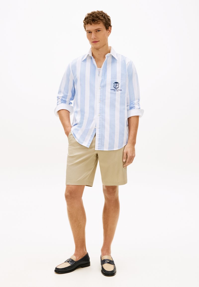 Άνδρας που φοράει μπλε και λευκό πουκάμισο με κάθετες ρίγες, μπεζ σορτς και δίχρωμα μοκασίνια, στέκεται με τα χέρια μερικώς στις τσέπες.