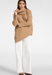 Kobieta ubrana w oversize'owy karmelowy sweter z dużym splotem i asymetrycznym dołem, białe szerokie spodnie oraz ciemnobrązowe klapki na obcasie.