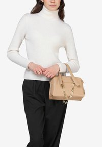 Borsa a tracolla in pelle beige con rifiniture dorate, manici superiori e una catena removibile. Indossata da un modello in un dolcevita bianco a coste e pantaloni neri.