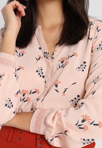 Blusa rosa chiaro con motivi floreali in corallo e blu navy. Presenta un design con bottoni, maniche arricciate e polsini scintillanti.