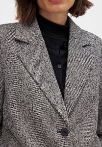 Blazer s čierno-bielym vzorom houndstooth, s výstrihom do "V", jedným čiernym gombíkom a textúrovaným materiálom, cez čierny rolák.