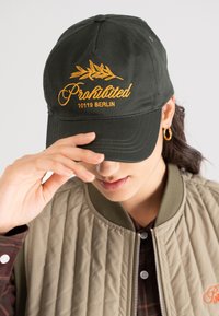 Person wearing a dark green cap with yellow text "Prohibited 10119 BERLIN," beige quilted jacket, and gold hoop earrings, adjusting the cap brim.