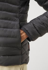 Giacca nera isolante con design trapuntato, dotata di tasca con zip, polsini elastici e etichetta con logo. Il materiale sembra leggero e liscio.