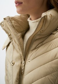 Beige pufferjack met een hoge kraag, horizontaal gestikte textuur, gouden hardware en een ritssluiting aan de voorkant. Draagt over een crèmekleurige trui.