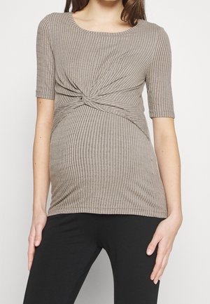 Gravid person iført en beige ribbet, tvistet front graviditetstop og sorte leggings, stående foran en ensfarvet baggrund.