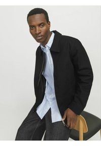 Veste noire avec fermeture éclair, col et poches latérales portée par-dessus une chemise à rayures bleu clair et blanche. Pantalons en denim foncé.
