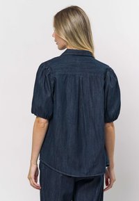 Donkerblauwe spijkershirt met schuine korte mouwen, een klassieke kraag en een licht gebogen zoom. De stof heeft een gladde textuur en een gemiddeld gewicht.