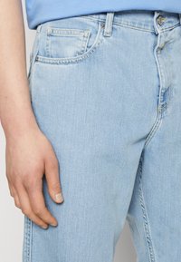 Pantalones vaqueros de mezclilla azul claro con un diseño clásico de cinco bolsillos. Presentan costuras visibles, una cremallera en la bragueta y una textura ligeramente desgastada.