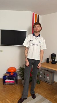 Balti polo marškinėliai su mėlynu apykakliu ir atspausdintu logotipu, derinami su pilkais džinsais ir bordo spalvos batais. Fone matomas televizorius ir spalvinga siena.