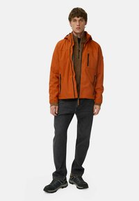 Orange waterproof jacket with adjustable hood, zippered pockets, layered over a brown patterned sweater, paired with black jeans and sturdy shoes.