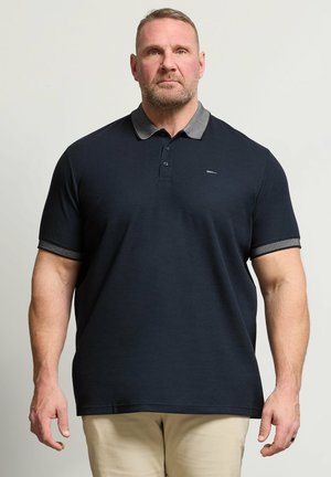 Mand med kort hår og skæg iført mørkeblå poloshirt og beige bukser, stående mod en ensfarvet lys baggrund.