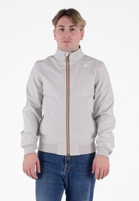 Giacca zip grigia chiara con colletto alto, dettagli a strisce arancioni e gialle, e polsini a costina, abbinata a jeans blu.