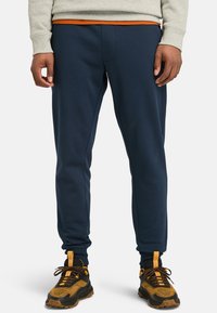 Navy blue sweatpants with elasticized waistband and cuffs, made from a soft fabric. Paired with black and orange patterned sneakers.