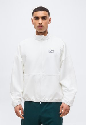 Νέος άνδρας που φοράει λευκό μπουφάν EA7 Emporio Armani με φερμουάρ και σκούρο πράσινο παντελόνι, στέκεται μπροστά σε ένα απλό ανοιχτόχρωμο φόντο.