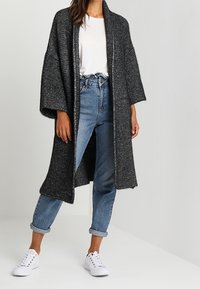 Svart och vit mönstrad lång kofta med en lös passform, vida ärmar och mjuk textur, lagd över en vit topp och blå jeans.