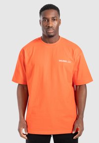 Př oversized oranžovém tričku z bavlny s kulatým výstřihem a krátkými rukávy. Na levé straně hrudníku se nachází bílé logo.