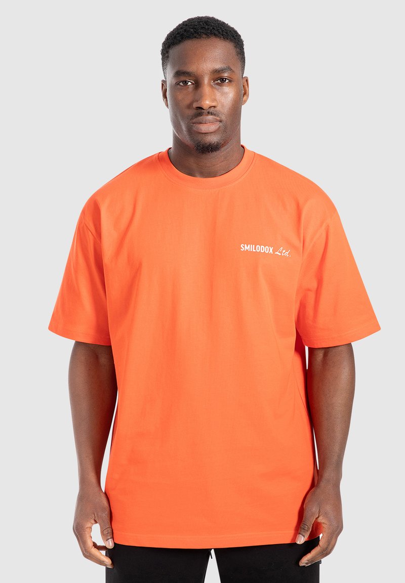 Př oversized oranžovém tričku z bavlny s kulatým výstřihem a krátkými rukávy. Na levé straně hrudníku se nachází bílé logo.