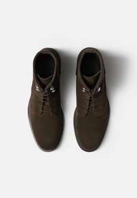 Botas de tobillo de ante marrón con puntera redondeada, seis ojales y detalles de hardware metálico. Suelas de goma planas y textura suave.