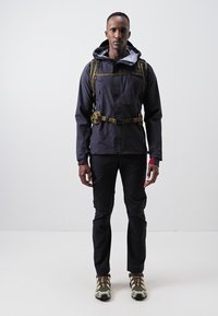 Water-resistant black jacket with a grey hood, featuring side zippers, worn over black pants and green-accented shoes, with a backpack.