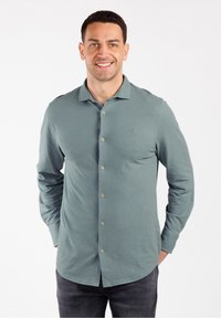 Uomo sorridente che indossa una camicia a maniche lunghe, con bottoni di colore teal con un piccolo logo sul petto e pantaloni scuri, in piedi davanti a uno sfondo bianco.