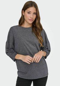 Mujer con largo cabello castaño ondulado que lleva una blusa gris suelta de manga larga brillante y pantalones negros, de pie con las manos entrelazadas.