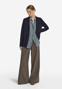 Marineblazer über einem hellblauen gemusterten Hemd, kombiniert mit weiten braunen Hosen und passenden Schuhen, was einen geschichteten, maßgeschneiderten Look kreiert.