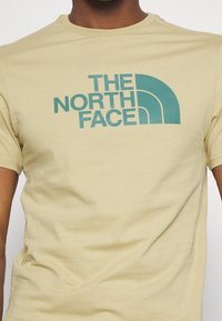 Camiseta de algodón beige con un logo turquesa que incluye un diseño de arco, con mangas cortas y un escote redondo.