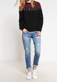Felpa nera con motivo camouflage rosso e testo "VERSUS"; abbinata a jeans chiari strappati e sneakers bianche.