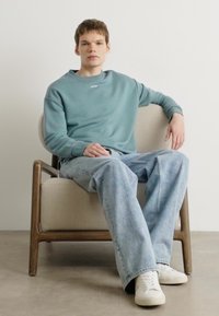 Sudadera azul clara con cuello de canalé, corte relajado, combinada con vaqueros anchos de denim claro y zapatillas blancas, sentado en una silla de madera.