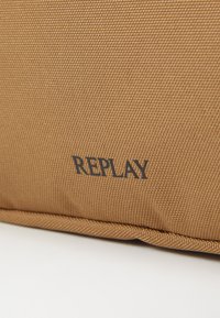 Cuscino marrone con texture e un motivo geometrico. Il marchio "REPLAY" è stampato in blu scuro su un lato.