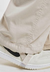 Pantaloni in cotone beige con orlo cucito e vestibilità morbida, abbinati a sneakers bianche e beige chiaro con un baffo nero e cintura.