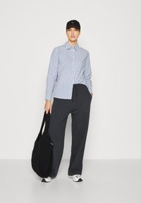 Camicia a strisce blu e bianche con bottoni, cappellino nero, pantaloni grigi con cintura a coulisse e borsa a rete nera. Il modello indossa sneakers bianche.