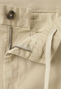 Beżowa talia spodni z guzikiem, częściowo otwartym metalowym zamkiem błyskawicznym i białym plecionym sznurkiem przewleczonym przez szlufkę paska.