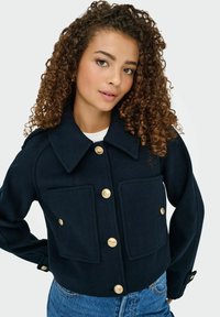 Marineblå uldjakke med struktureret krave, der har store frontlommer og guldtonede knapper. Designet er cropped og tætsiddende ved taljen.