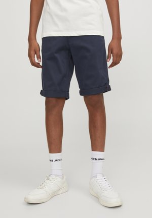 Person iført marineblå shorts med opsmøg, hvide crew-sokker med sort tekst, hvide sneakers og en hvid t-shirt, stående mod en ensfarvet baggrund.