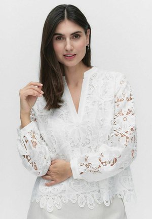 Vit blus med blommönster i spets, långa ärmar och V-hals. Den har intrikat broderi och en vågig nederkant.
