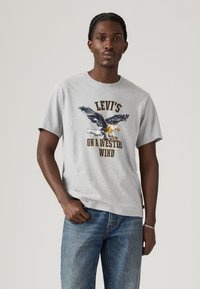 Man met een lichtgrijze Levi's t-shirt met een adelaar graphic en de tekst "On a Western Wind", gecombineerd met blauwe jeans en een zilveren armband.