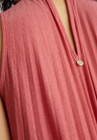 Primo piano di un indumento rosa texture senza maniche con scollo a V e un piccolo ciondolo metallico rotondo al colletto.