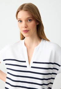 Junge Frau mit glattem blonden Haar und blauen Augen, die ein weißes Hemd mit horizontalen marineblauen Streifen und einem V-Ausschnitt trägt.