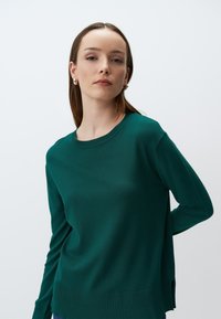 Zielony sweter dzianinowy z okrągłym dekoltem, długimi rękawami, bocznymi rozcięciami i gładką fakturą. Materiał wygląda na miękki i lekki.