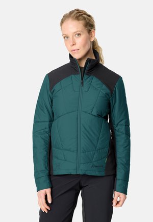 Frau trägt eine türkis-schwarze isolierte Jacke mit Reißverschluss und hohem Kragen, steht vor einem schlichten hellen Hintergrund.