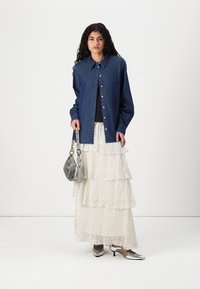 Denimskjorta i mörkblått med ficka, lager av vit prickig nederdel, silvriga klackskor och en metallisk handväska. Enfärgad bakgrund.