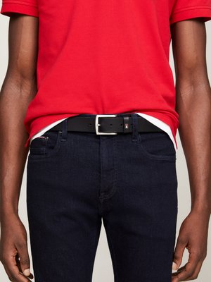 Cintura in pelle nera con fibbia argentata, indossata sopra jeans blu scuri. La polo rossa è visibile all'altezza della vita, con una maglietta bianca sotto.