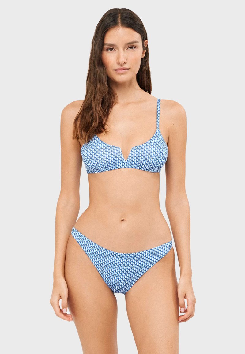 Femme portant un bikini bleu et blanc à motif avec de fines bretelles et une petite encoche en forme de V sur le haut, debout sur un fond clair uni.