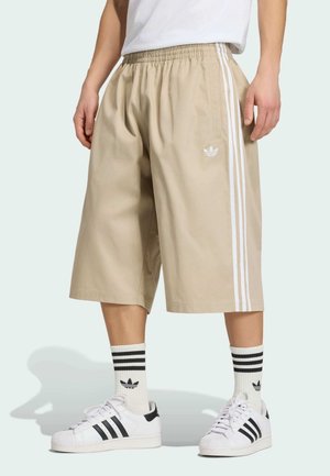 Beżowe szorty o szerokich nogawkach z białymi bocznymi pasami i logo, w połączeniu z białymi butami Adidas oraz białymi skarpetkami z czarnymi paskami.