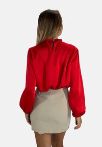 Blusa rossa in satin con maniche a sbuffo e dettaglio a chiave sulla schiena, abbinata a una mini gonna fitted beige. Texture liscia, colore vivace.