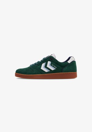 Groene suède sneaker met witte accenten, ronde neus, donkerblauwe hiel, rubberen zool en contrasterende diagonale strepen aan de zijkant.