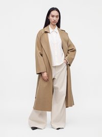 Femme portant un long manteau couleur camel, une chemise blanche à boutons, un pantalon beige large et des chaussures noires, debout avec une main dans la poche sur fond blanc.
